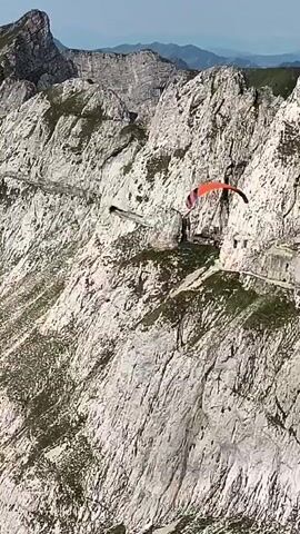 A paraglider in an orange and red wing is soaring near a sheer cliff face. The paraglider is positioned mid-air, with the cliff face to its right. Below the paraglider, a dark, elongated object, possibly a drone or another paraglider, is visible. The cliff face itself is rugged and textured, with patches of green vegetation clinging to its surface. In the background, jagged mountain peaks rise against a hazy sky. A narrow pathway or road is discernible carved into the side of the mountain, running along the cliff.