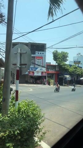 The video shows a daytime street view from inside a moving vehicle. The street is lined with various buildings, including coffee shops, car service shops, and sport stores. Vehicles, including motorcycles and cars, can be seen driving along the road. There are also trees and shrubs planted along the roadside. The sky is clear and blue. A banner advertising "NAM DU LICH QUOC GIA LAI 2026" is visible on a light pole.