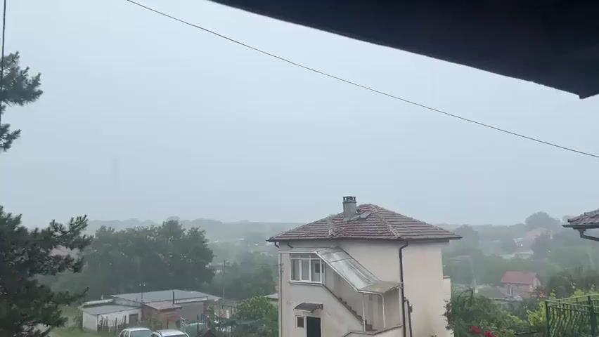 The video presents a static, elevated view of a residential area in Dimchevo, Bulgaria, during a heavy downpour. The sky is entirely overcast and grey, creating a subdued and misty atmosphere.

Heavy rain is visibly falling throughout the scene, appearing as streaks across the frame and creating splashes on visible surfaces. The intensity of the rain contributes to a hazy effect in the background.

In the midground, a prominent two-story house with a reddish-brown tiled roof and a light-colored facade stands out. It features an external staircase leading to an enclosed upper-level balcony or sunroom, and a chimney is visible on its roof. To the left, several cars are parked near a cluster of smaller buildings. Beyond these, the landscape is dotted with more houses and abundant green trees and foliage, fading into the distance due to the rain and mist. A thin wire or cable stretches diagonally across the upper portion of the frame, close to the viewer.

No people are visible, and the primary activity is the continuous heavy rainfall. The time of day is daylight, but the severe weather conditions make the scene appear dark. There is no visible text in the video.