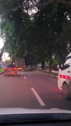 The video captures a first-person perspective from inside a vehicle driving on a multi-lane road at dusk or early evening in Siron, Indonesia. The road is lined with abundant green trees on both sides, creating a somewhat shaded canopy. The sky, visible in the distance, is bright with an overcast appearance.

Ahead of the viewer's vehicle, a dark-colored SUV is visible, its taillights illuminated. Behind the SUV is a large, light-colored truck with its headlights on. To the right of these vehicles, a white ambulance van with red and white stripe markings is prominent. As the video progresses, a person on a motorcycle briefly appears, passing the ambulance on the right side.

The ambulance's rear features the word "AMBULANCE" and a clear message "SAVE PALESTINE" above a graphic. Vehicle lights are active, including the taillights of the SUV and ambulance, and the headlights of the truck. Streetlights are also visible along the right side of the road, contributing to the evening ambiance. The scene depicts typical traffic flow during the transition from day to night.