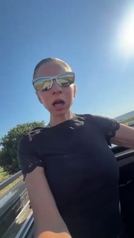 A woman with blonde hair pulled back and wearing sunglasses and a black t-shirt leans out of a vehicle. She is looking at the camera with her mouth open. The sun is shining brightly in a clear blue sky. The vehicle appears to be in motion, with a landscape of dry grass and hills visible in the background.