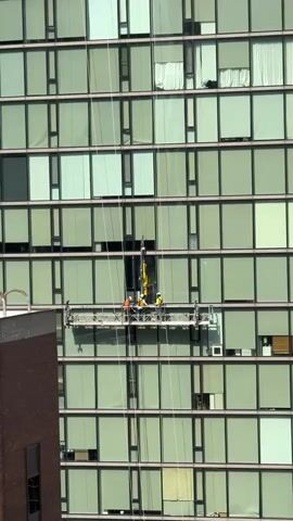 Workers are performing maintenance on a high-rise building. A suspended platform, commonly known as a swing stage, is attached to the building by cables and is being used by several individuals. The platform is positioned mid-way up the building, which has a facade of green glass windows. The workers are wearing safety vests, indicating they are engaged in construction or repair work. The overall scene depicts a typical urban environment with ongoing construction or maintenance activities at considerable height.