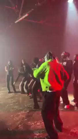 The video captures a group of people dancing in a dimly lit, industrial-style space, possibly a warehouse. Red lights and fog create a hazy atmosphere. Most of the dancers are dressed in dark clothing, while one person stands out wearing a bright green jacket. Some of the individuals are facing away from the camera. A person in a blue and black jacket appears to join in the dance briefly. The space has a concrete floor with visible marks and streaks. The background shows exposed ceiling beams and ductwork, characteristic of an industrial setting.