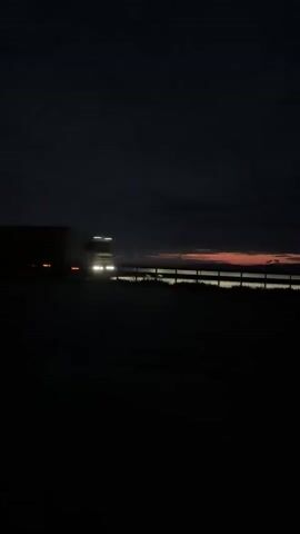 The video presents two distinct nocturnal or low-light scenes in Cherna Gora, Bulgaria.

The initial frames capture a semi-truck traveling on a dark road during what appears to be either dawn or dusk. The sky above is dark, with a subtle horizontal band of orange and red color along the horizon, hinting at the rising or setting sun. The truck, a large tractor-trailer, is a dark silhouette against the faint sky, moving from left to right across the frame. Its speed is indicated by motion blur, and a few amber side marker lights are faintly visible on its trailer. After the truck passes, a streak of red light, likely its taillights, briefly lingers on the right side of the screen.

The video then transitions to a different setting, clearly at night. A dark-colored sedan, possibly a wagon, is shown in a paved area, which could be a parking lot or a side road. Its bright headlights cut through the darkness, illuminating the ground directly in front of the vehicle. To the left, a brightly lit rectangular object, possibly a container or a small structure, is visible, along with some green vegetation beyond it. The car appears to be stationary or moving very slowly in this unlit environment. No people or visible text are present in the video.