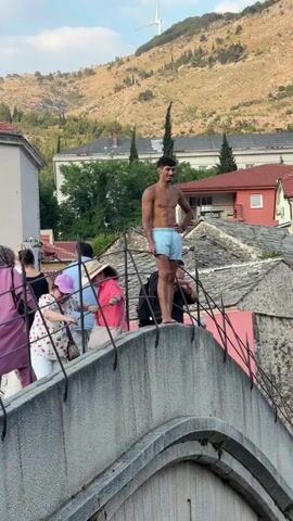 The media file captures a scene on the Stari Most (Old Bridge) in Mostar, Bosnia and Herzegovina. The iconic stone bridge, with its distinctive arch, spans a deep, turquoise river. Several people are gathered on the bridge. In the foreground, a young man in light blue shorts stands shirtless, appearing poised. He is surrounded by other individuals, some of whom are tourists observing the scene.

Behind the bridge, traditional Ottoman-era buildings with red-tiled roofs and stone facades line the riverbanks. Lush green trees and vegetation climb the hillsides in the background, where a wind turbine is visible on the crest of a mountain. The sky is clear and bright, suggesting a pleasant day, likely late afternoon or early evening, given the warm light.

The prominent activity suggested by the context of the Stari Most is the tradition of young men diving from the bridge into the Neretva River below, a practice that draws crowds and tourists. While the man in the foreground is not actively diving in this particular frame, his stance and the onlookers suggest he may be preparing to or has just finished a dive. The overall atmosphere appears to be one of excitement and cultural appreciation, with tourists eager to witness this local tradition. The setting is distinctly historic and picturesque.