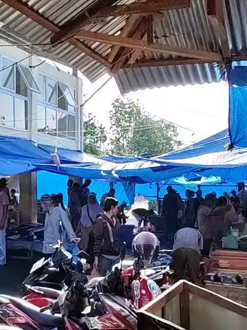 The video shows a bustling outdoor market.  People are milling about, some wearing traditional Muslim clothing, browsing vendors' tables under blue tarps.  Motorcycles are parked throughout the market area. Some individuals are standing, while others are seated on low stools or benches. Products for sale appear to include fresh seafood, likely fish, given the proximity to the ocean. The market is sheltered by a corrugated metal roof supported by white pillars. A two-story building stands in the background with multiple windows. The ground is wet and appears to be made of concrete or stone.