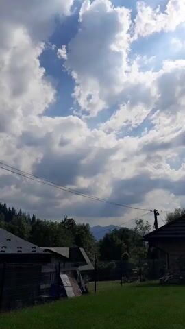 The media file depicts an outdoor scene in Gliczarów Górny, Poland. The sky is filled with dramatic, puffy white clouds against a bright blue backdrop, suggesting it is daytime, likely mid-morning or early afternoon, with sunny intervals. The weather appears partly cloudy and fair.

The scene looks like a rural or semi-rural residential area. In the foreground, a well-maintained green lawn is visible. Beyond the lawn, there are houses with dark, sloped roofs, some appearing to be made of wood, with visible construction elements like a ladder leading to a higher level of one building. A black wire fence encloses part of the property. Power lines stretch across the mid-ground, supported by a utility pole.

Behind the houses and trees, a line of distant mountains can be seen, their peaks softened by the atmospheric haze. The vegetation consists of lush green trees, forming a dense treeline that borders the residential area. The overall impression is one of a peaceful, scenic landscape, typical of a mountainous or hilly region. There is no visible text in the media file, nor are there any people or animals. The activity taking place is simply the static view of the environment. The emotion evoked is one of tranquility and natural beauty.