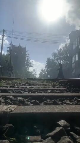 The video shows a low-angle, eye-level shot of a railroad track with dense foliage and buildings on either side. The sun is high in the sky, suggesting it is daytime, and the bright light creates a slight haze. Palm trees are visible, hinting at a tropical location, which is further confirmed by the context of Kodagoda, Sri Lanka.

In the background, several people are engaged in different activities. A person is walking across the tracks, while another is riding a bicycle. A motorcycle is also seen moving along the path beside the tracks. The overall atmosphere appears to be peaceful and ordinary, depicting a typical day in a populated area with a railway line. The mood is serene, with the natural light playing a significant role in the visual aesthetic. There is no visible text in the video.