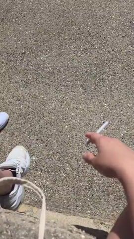 The video begins with a close-up shot of a hand holding a cigarette against a grey, pebbly ground. The camera then pans up and away, revealing a wider scene on a bright, sunny day. In the background, several people are gathered, some standing, some sitting, and others on bicycles. A few individuals are walking or cycling on an asphalt path. The environment appears to be outdoors, possibly near a beach or a recreational area, indicated by the presence of sand and the clear blue sky. The focus shifts to a young person riding a green bicycle, who is the central figure in the latter half of the video. Other people are visible in the distance, scattered across the scene. The overall atmosphere suggests a casual, outdoor activity.