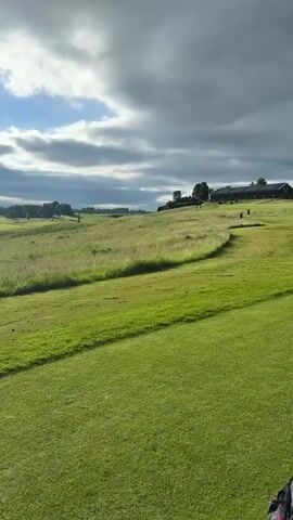 The video pans across a lush green golf course under a cloudy sky. In the distance, several golfers can be seen playing, and a few golf carts are also visible. The landscape is rolling, with trees and fields in the background. A body of water, possibly a lake or pond, is also visible in the distance. The sun intermittently breaks through the clouds, casting beams of light across the course. The overall impression is one of a peaceful and scenic golfing environment.