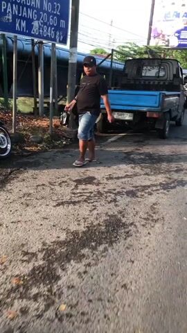 This is a short video clip, appearing to be taken from a moving vehicle, showing a man walking along a roadside in Ngaban, East Java. The man is wearing a black t-shirt, denim shorts, a baseball cap, and flip-flops. He is carrying a dark bag in his left hand. He walks from left to right across the frame.

Behind the man, a small blue pickup truck is parked. The truck has a metal cage on its bed. To the left of the man and the truck is a blue signpost with white text and some symbols. The text on the sign is partially visible and reads "NGABAN NO.32 PANJANG 20,60 m KM. 20+846". This suggests the man is walking along a road in the Ngaban area of East Java, and the sign may be indicating a road number and length.

The ground is paved but has patches of dark, wet-looking marks, possibly from oil or water. There are also some fallen leaves on the ground near the signpost and the truck. Sunlight is casting shadows, indicating it is likely daytime, possibly late afternoon or early morning due to the angle of the light. The overall atmosphere appears to be casual and everyday. No strong emotions are conveyed by the subject. A blurry advertisement billboard is visible in the background above the truck.