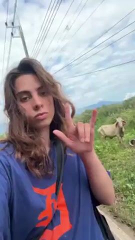 A young person with brown hair and wearing a blue t-shirt with an orange design makes the "I love you" hand gesture in front of the camera.  A cow is visible in the background, grazing in a grassy area.  Power lines stretch across the sky above.  The scene appears to be outdoors, possibly on a road or pathway with vegetation on one side. The person appears to be taking a selfie video.