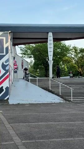 A person wearing a black t-shirt, dark baggy pants, and a white helmet is captured performing a scooter trick in an outdoor urban setting in Saint-Priest, France. The individual starts on a slightly elevated concrete ramp, then jumps onto a white metal handrail, grinding down its length alongside a large concrete staircase. After a successful slide, they land smoothly on the paved ground below and ride away.

The environment features modern architecture with a mix of concrete and metal structures. To the left, a building wall is decorated with a large, abstract mural composed of red, white, and black geometric shapes. In the background, two prominent, tall, white decorative pillars, shaped like elongated fins, stand symmetrically. Behind the staircase, several other people are casually present, some sitting on the steps, others standing further back near lush green trees.

The scene appears to be taking place during the daytime, likely late afternoon or early evening, characterized by soft, clear lighting and a bright sky. A large, dark overhead structure, possibly a bridge or an extended roof canopy, frames the top portion of the view. In the distance, a multi-story residential building is visible. There is no clearly legible text in the video.