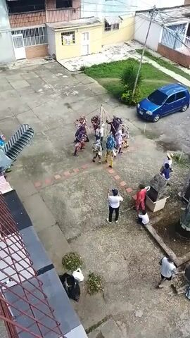 This video shows an outdoor scene in what appears to be a courtyard or open paved area, possibly a school or community space. In the center of the frame, a group of people, likely children or young adults, are engaged in a performance or dance. They are dressed in colorful and elaborate costumes, some adorned with feathers and intricate patterns. The group is gathered around a central structure, possibly a decorated pole or effigy, which they are interacting with, suggesting a ritual or celebratory event.

Several other individuals are observing the performance, standing or sitting around the edges of the paved area. One person in a white shirt and black pants is holding a phone, seemingly recording the event. Another person in a red shirt is also watching, and a child in white sits nearby. Further to the right, another group of observers is visible, including a person in a blue shirt and another in white. A blue minivan is parked in the background. The setting includes buildings with balconies and windows, some with awnings, and patches of grass and greenery. The ground is paved with concrete.

The lighting suggests it is daytime, with a moderate amount of light, but the sky is not clearly visible to determine the exact weather. The overall atmosphere appears to be one of community gathering and cultural expression. There is no discernible text in the video. The presence of costumes and the organized performance strongly suggest a cultural celebration or festival taking place in Tinaquillo, Venezuela.