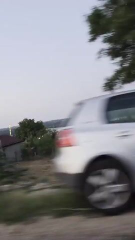The video appears to be recorded from a moving vehicle, likely a car, looking out the side window. The camera is tilted upwards and to the side. In the foreground, there is a blurred landscape of grass and trees. There are glimpses of a car's side mirror, showing a reflection of the interior of the car, including what seems to be a person's arm. In the mid-ground, there are some houses with reddish-brown roofs and other structures that are difficult to discern due to motion blur. Beyond the houses, there are more trees and what appears to be a body of water, possibly a river or a lake, in the distance. The sky is overcast with a pale, muted color, suggesting it might be early morning or late evening. The overall impression is a rural or semi-rural setting observed during a journey.