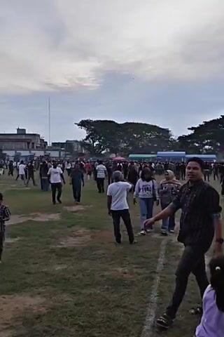 The video shows an outdoor gathering of people in a grassy field. There are several tents set up in the background, suggesting some sort of event or market. A large, leafy tree dominates the middle ground. The crowd appears to be diverse, with men, women, and children present, many of whom are wearing headscarves. The sky is overcast. People are walking around and interacting with each other. The camera is handheld and appears to be capturing footage from within the crowd.
