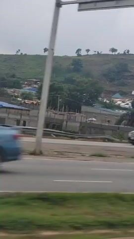 The video shows a roadside view of a town built on a hill. A highway is in the foreground, with a concrete barrier and green grass lining its edge. Beyond the highway, there are buildings under construction, and many houses with corrugated iron roofs are scattered across the hillside. Several vehicles are parked on the side of the road, and some people can be seen in the distance. The sky is overcast. The camera appears to be moving, suggesting the video was filmed from a vehicle.