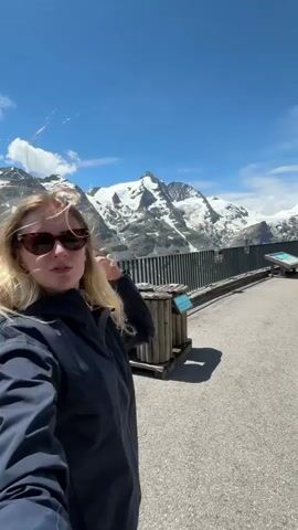 A blonde woman wearing sunglasses and a dark jacket takes a selfie outdoors with snow-capped mountains in the background. She is standing on a paved area with a wooden fence and some informational signs. There are a few other people in the background, including a man in a white t-shirt and shorts. The sky is blue with a few clouds.