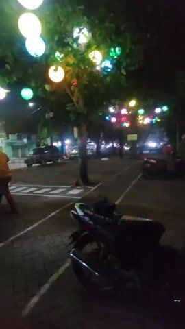 Here is a concise description of the video:

The video captures a nighttime scene on a city street.  A tree, decorated with colorful spherical lights, dominates the foreground.  Behind the tree, a figure walks in the distance on the sidewalk.  A scooter is parked near the tree.  A building with bright lights and an arched facade is visible across the street. The overall lighting is dim, with the colored lights and building illumination being the primary sources of light. The focus is blurry.