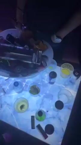 The video shows a nightclub scene. A marble-patterned table is illuminated with blue light. On the table are numerous glasses, cans, and bottles, along with a large ice bucket filled with more drinks and bottles. Someone's hand reaches into the ice bucket. The camera pans up to show a crowd of people in a dark room, with dynamic geometric lighting displays in blue and pink. Music plays in the background.