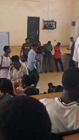 Here's a description of the video:

The video shows a room filled with people, mostly students, in what appears to be a classroom or lecture hall. Some individuals are standing and moving around, while others are seated at desks. The room has cream-colored walls, a whiteboard, and a dark-colored door. The people appear to be socializing and walking about. The camera is positioned among the seated students, looking towards the front of the room where most of the activity is taking place.