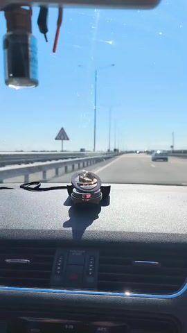 A first-person perspective video shows the interior of a car driving on a highway. The camera is mounted on the dashboard, facing forward. A car air freshener hangs from the rearview mirror, and a circular electronic device, possibly a compass or a speed gauge, sits on the dashboard in front of the passenger seat. The highway is lined with barriers and streetlights, and other vehicles can be seen in the distance. The sky is clear and blue. The video is shaky, indicating the car is in motion.