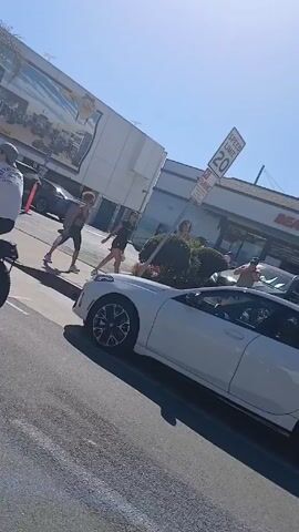 The video shows a street scene with several people walking near a white BMW parked on the side of the road. The car is parked in front of a business called "Beach Market," which features a sign with the words "Hermosa Beach." There is a speed limit sign indicating 20 mph. Several other cars are parked in the vicinity. The sky is clear and blue. The video appears to be taken from a moving vehicle, possibly another car or a bike.