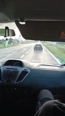 The video shows the interior of a vehicle from the passenger's perspective. The dashboard and the driver's seat are visible in the foreground. The view through the windshield reveals a road with a black SUV in front. The road appears to be a two-lane highway with white markings. The weather seems to be overcast, and there is vegetation along the sides of the road. A sign with a red exclamation mark inside a circle is visible on the passenger side visor. The car is in motion, following the SUV.