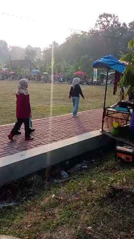 Two young girls are walking on a brick pathway. The girl on the left is wearing a red jacket and a hijab. She is holding a green object. The girl on the right is wearing a blue jacket and a hijab. They are walking away from the camera. In the background, there is a grassy field with some tents and umbrellas. The sky is bright and hazy. There are some trees in the distance.