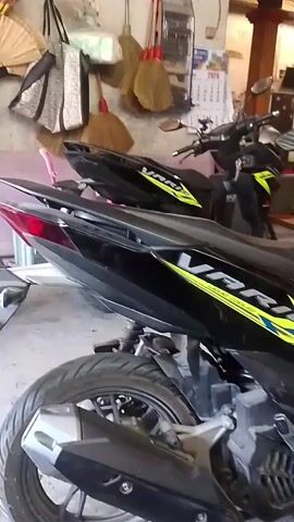 The video shows two black scooters parked in what appears to be a garage or storage area. The scooters have yellow and white accents. One scooter is in the foreground and is mostly visible from the rear, showing its exhaust and rear tire. The second scooter is behind the first and is more visible from the side, showing its side panel with the word "VARIO" written on it in yellow and blue. In the background, there are various items hanging on the wall, including what look like brooms and bags, as well as a calendar. A helmet is placed on the handlebars of the second scooter. The floor is concrete and appears somewhat dirty. The lighting suggests an indoor environment, possibly with natural light coming from a window or opening.