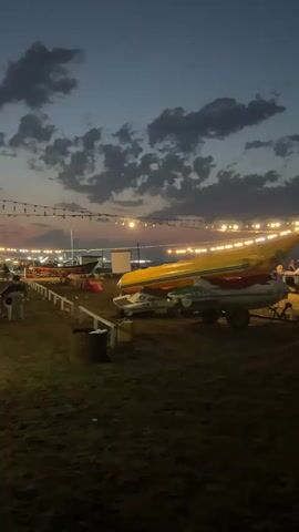 The video depicts a beach scene at dusk. Several jet skis and boats are parked on the sand, some on trailers. String lights are visible overhead, illuminating the area. In the background, the sea is visible, and the sky is a deep blue with scattered clouds. There is a lifeguard tower and a small structure on the beach. A few people are present, though their actions are not clearly discernible due to the low light. The overall atmosphere is calm and serene, suggesting the end of a day at the beach.