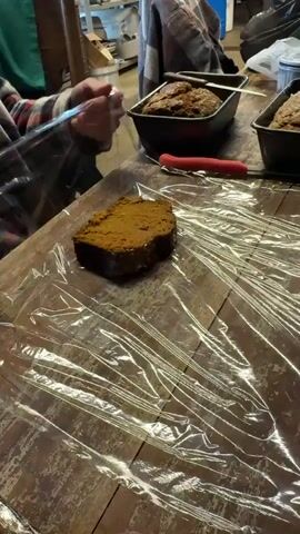 A person in a red and gray plaid shirt is seen wrapping a loaf of bread in plastic wrap. The person is sitting at a wooden table. There are two bread pans in the background, both containing baked goods. A red-handled knife sits near the bread pans. The environment appears to be inside a rustic-looking building, possibly a farmhouse or workshop. There are shelves and other items in the background, slightly out of focus. The person is handling the plastic wrap carefully, ensuring the bread is fully covered. The video focuses on the action of wrapping the bread.