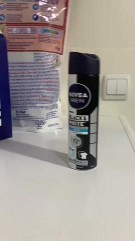 The video shows a close-up shot of a Nivea Men Black & White deodorant spray can. The can is black and white with blue accents and has the Nivea Men logo. The can is standing upright on a white textured surface, which appears to be the top of a washing machine or dryer. To the left of the deodorant can, a portion of a blue box is visible, and behind the deodorant can, a pink bag with text and images, possibly laundry detergent, is also visible. To the right of the deodorant can, a white light switch with two toggles is mounted on a white tiled wall. The lighting is bright, suggesting an indoor setting. The camera slowly zooms in and out, providing a detailed view of the deodorant can and its surroundings.