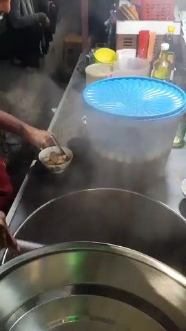 A person's hands are visible, using tongs to place food into a bowl from a large pot. The pot is steaming. To the right of the pot is a large clear plastic container with a blue lid, filled with brown liquid and small round objects. A person's arm is visible to the left, leaning over the counter, and another person is partially visible in the background, seated. Various containers and bottles are on a shelf behind the counter. The setting appears to be an outdoor food stall at night.