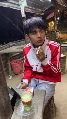The video shows a young person sitting at a table outdoors at night. They are wearing a red and white jacket and jeans, and they are holding a cigarette to their lips. In front of them on the table is a glass with a layered drink, a pack of cigarettes, and a tablet device. In the background, there are lights and foliage, suggesting a somewhat rural or natural setting. The person appears to be looking at the camera or something just off-camera. The overall atmosphere is casual and possibly a bit secluded.