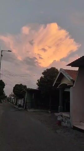 The video shows a rural street scene at dusk. A large, fluffy cloud glows with orange and pink hues from the setting sun. The street is paved with asphalt and lined with houses and trees. Power lines stretch across the sky. The perspective is low, looking up towards the sky and down the street.