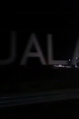 A shaky video shot at night shows a large, illuminated sign. The sign consists of individual, white, three-dimensional letters against a dark background. The letters appear to spell out "TOLAN". Some of the letters are out of focus or partially obscured by motion blur. In the background, faint lights of what appears to be a city or industrial area are visible. The foreground shows a blurred dark surface, possibly a road or barrier, suggesting the video is taken from a moving vehicle.