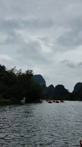 The video shows a river scene on a cloudy day. Several small boats, each carrying people wearing orange life jackets, are on the water. The boats appear to be moving slowly, possibly being rowed. The river is surrounded by lush green vegetation and tall, rocky hills or mountains.  The water is dark and choppy, reflecting the overcast sky. In the foreground, a small portion of the videographer's head and a red flag are visible.