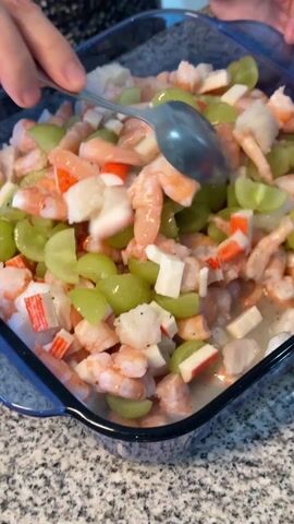 A person stirs a seafood salad in a blue glass square bowl with a silver spoon on a gray speckled countertop. The salad is comprised of small shrimp, green grapes cut in half, white fish cubes, and imitation crab cut into small pieces. The person's hand is partially visible in the frame. They are wearing a black and white patterned shirt.