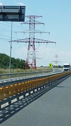 The video shows a view from a vehicle moving along a highway. There are large electrical transmission towers visible on the right side of the highway. A truck with a tank trailer is seen in the distance on the highway. Road signs indicating destinations such as Bucharest, Cartier, Rahova, and Bragadiru are visible. A silver sedan is in front of the camera vehicle on the highway. The weather appears to be clear and sunny. The highway has multiple lanes and is divided by a barrier with yellow reflective elements. Trees and vegetation are visible on the sides of the highway.