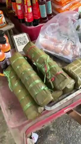 The video shows a close-up of a metal tray filled with various packages wrapped in banana leaves. Some of the packages are long and cylindrical, tied with green and red plastic strings. Others are square-shaped, bound with thin, light-colored natural twine. A clear plastic bag containing what appear to be small, brown sausages rests next to the wrapped packages. In the background, blurred items like bottles and jars are visible on a shelf. A QR code is placed next to the cylindrical packages.