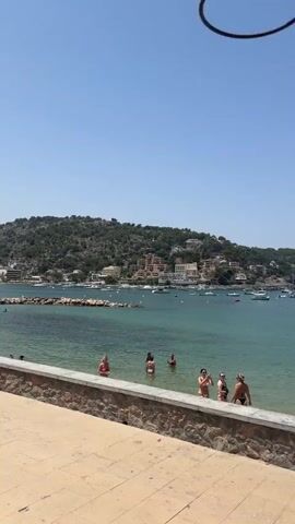 The video pans across a scenic bay on a clear, sunny day. In the foreground, a paved walkway runs along a low stone wall, separating it from a beach area. Several people are in the shallow water, some wading and others appearing to swim. Beyond the beach, the turquoise water of the bay is dotted with numerous boats of varying sizes, from small dinghies to larger yachts. In the distance, a picturesque town with white buildings and red roofs is nestled on a lush, green hillside that slopes down to the water. The sky is a vibrant, cloudless blue.