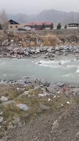 The video captures a view of a wide, rocky river with rapidly flowing water. The water appears slightly murky. In the background, there are some buildings and a mountain range obscured by a misty, overcast sky. On the riverbank, the ground is rocky and sparse vegetation is visible. A person wearing dark clothing walks across the river using a pole for balance. Further up on the bank near the buildings, there are two more people standing. A car and a van are also parked near the buildings.