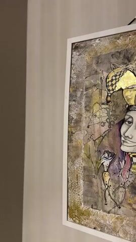 A close-up, side-angle shot of a framed artwork depicting a jester. The jester has a checkered hat and a slightly amused expression. The artwork itself is abstract and textured, with gold leaf accents and various mixed media elements. The background of the video is a plain, lightly textured wall. The lighting appears to be artificial and even. The video appears to be a still frame from a slow pan or zoom of the artwork.