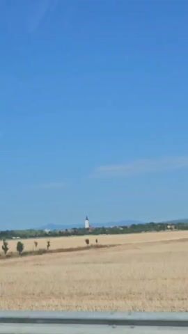 The video shows a view from a moving vehicle, possibly a car, on a sunny day. The landscape outside consists of a clear blue sky with a few wispy clouds. In the distance, there is a field of dry, golden-colored grass or stubble, possibly after a harvest. A small village with buildings and a church steeple is visible on the horizon, nestled amongst trees. To the left, there is a row of trees along a gentle slope. In the foreground, there is a metal barrier or railing. The vehicle passes under a concrete overpass, revealing a gray concrete embankment with steps on the side. The view then returns to the landscape outside. The overall impression is of a rural setting in fair weather.