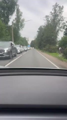 The video shows a view from inside a car looking out the windshield at a two-lane road.  There is a traffic jam with cars lined up in the left lane. Trees line both sides of the road. On the right side of the road, two people are standing on the shoulder. The sky is overcast. The car appears to be moving slowly forward.