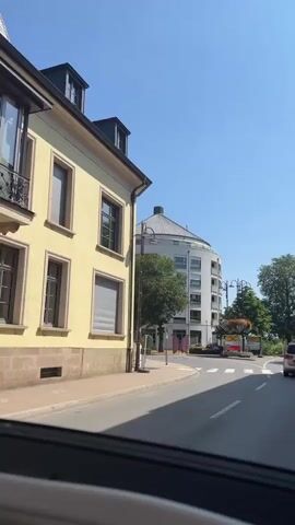 The video shows a street scene on a clear day. A yellow building is on the left, with a rounded white building further down the street. There are trees and streetlights visible. A car is driving on the road, and there are street signs. The perspective appears to be from inside a vehicle. There is a pedestrian crossing visible at the end of the video.