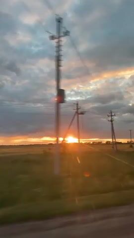 This media file captures a series of shots of a sunset in Рождествено, Russia. The primary subject is the dramatic sky, filled with a mix of dark, moody clouds and vibrant streaks of orange and yellow as the sun dips below the horizon.

The scene is an open, rural landscape, likely a field or grassland, bordered by trees and dotted with utility poles and power lines. The first shot shows a wide view of the sunset over the fields with utility poles framing the scene. Subsequent shots zoom in on the sky, highlighting the intense colors and cloud formations. One shot features an unusual lens flare or light phenomenon above the setting sun. The final shot shows the sunset partially obscured by blurred trees, suggesting movement, perhaps from a vehicle.

The time of day is clearly sunset, characterized by the warm, golden hues and lengthening shadows. The weather appears to be partly cloudy, contributing to the dramatic visual effect. The presence of utility poles and the open fields suggest a rural or semi-rural setting outside of a major urban center. The overall mood conveyed is one of natural beauty and perhaps a touch of mystery due to the unusual light effect and the slightly desolate landscape. No visible text is present in the media file.