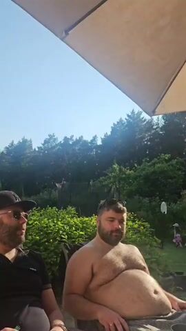 The video shows an outdoor gathering under a large umbrella. In the foreground, a man with a beard sits in a chair, looking towards the camera. To his right, a younger man stands, looking away from the camera. A table is set with food, including tomatoes, cucumbers, and what appears to be avocado salad, along with bottles of olive oil and spices. To the right, a woman in a purple dress is seated, cutting tomatoes. In the background, a woman with glasses and blonde hair takes a selfie. Further in the background, children are visible playing in a yard. The environment is a sunny day with trees and greenery surrounding the area.
