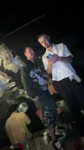 Two men are standing in a crowded, dimly lit room that appears to be a bar or club. The man on the left is wearing a black sweatshirt and shorts with high-top sneakers. The man on the right is wearing a white patterned short-sleeve shirt and black pants. Initially, the man on the right looks at his phone. He then looks up, smiles, and spreads his arms wide.  The two men turn around. The camera pans to show a wider view of the crowd, featuring various people socializing, some holding drinks. A decorative lighting fixture made of white material is visible above.